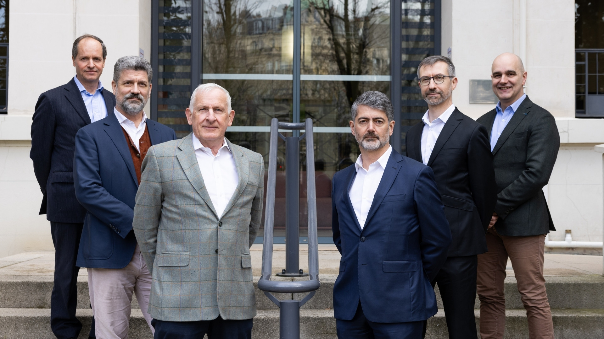 Six men in business attire standing on outdoor steps in front of a glass door and cream-colored building.