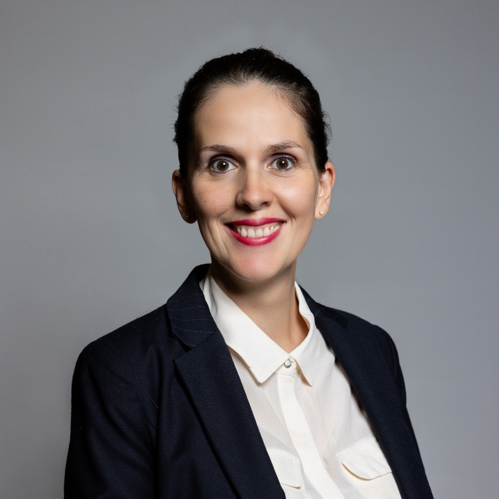 Smiling woman with dark hair pulled back wearing a white collared shirt and dark blazer against a gray background.
