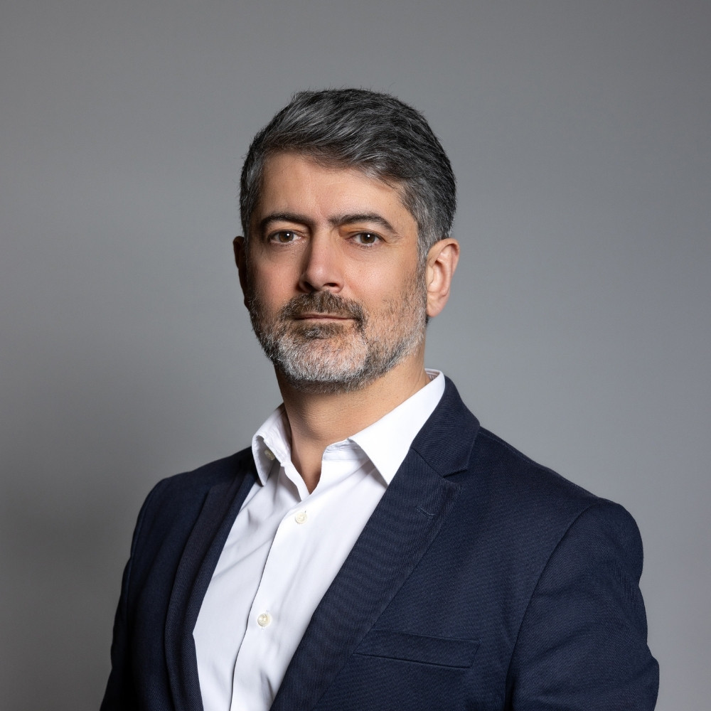 Middle-aged man with salt-and-pepper hair and beard wearing a navy blazer and white shirt against a gray background.