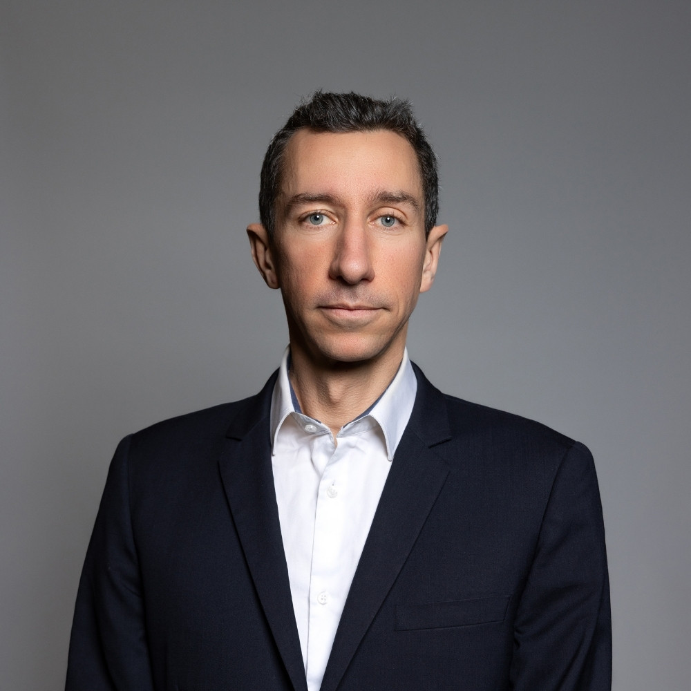 Man with short dark hair wearing a white shirt and dark blazer against a gray background.