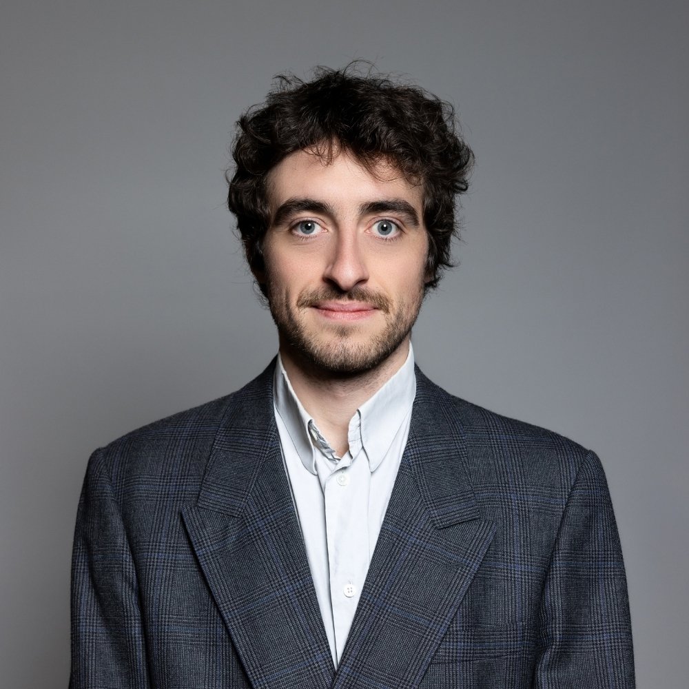 Man with curly brown hair and beard wearing a gray checkered blazer and white shirt against a gray background.
