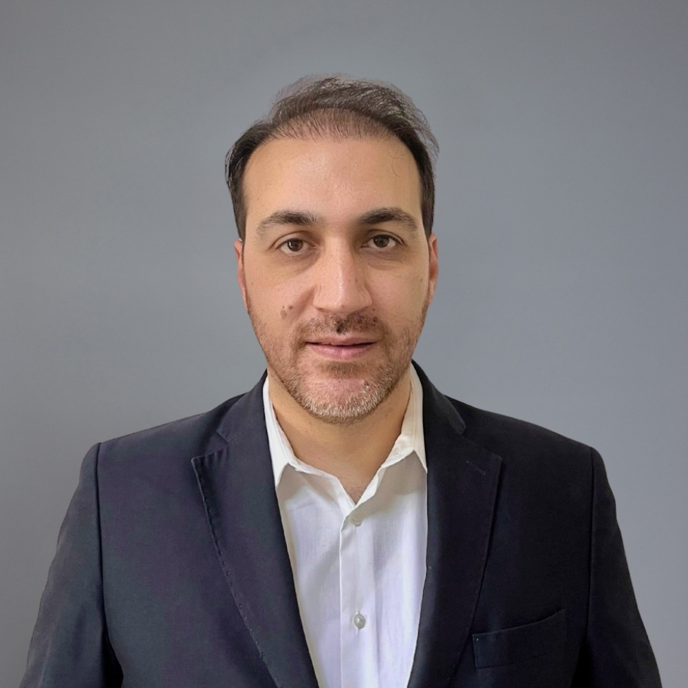 Middle-aged man with salt-and-pepper hair and beard wearing a navy blazer and white shirt against a gray background.