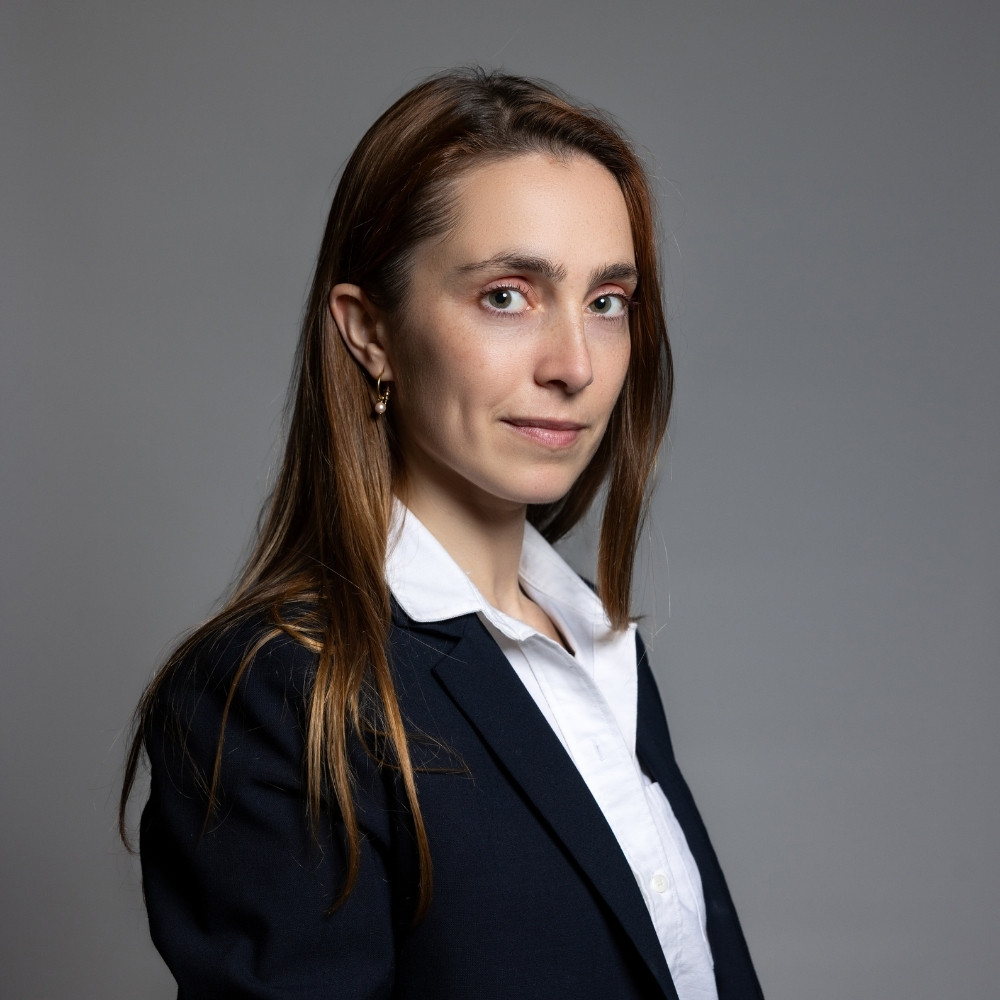 Professional woman with long brown hair wearing a white shirt and dark blazer against a gray background.