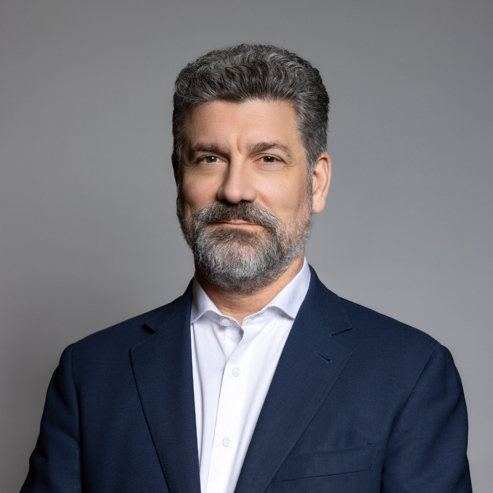 Middle-aged man with gray hair and beard wearing a white shirt and navy blazer against a gray background.