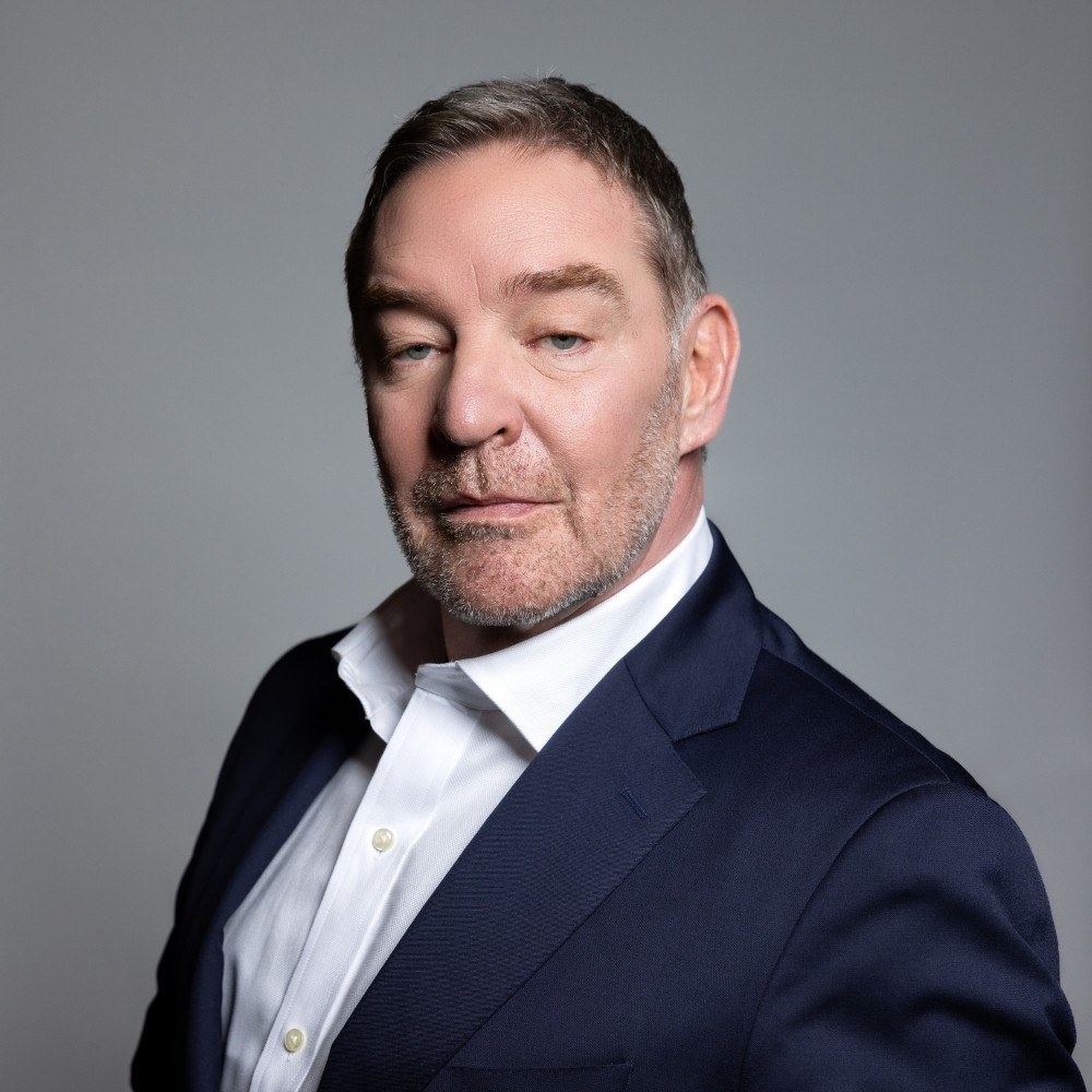 Professional woman with long brown hair wearing a white shirt and dark blazer against a gray background.