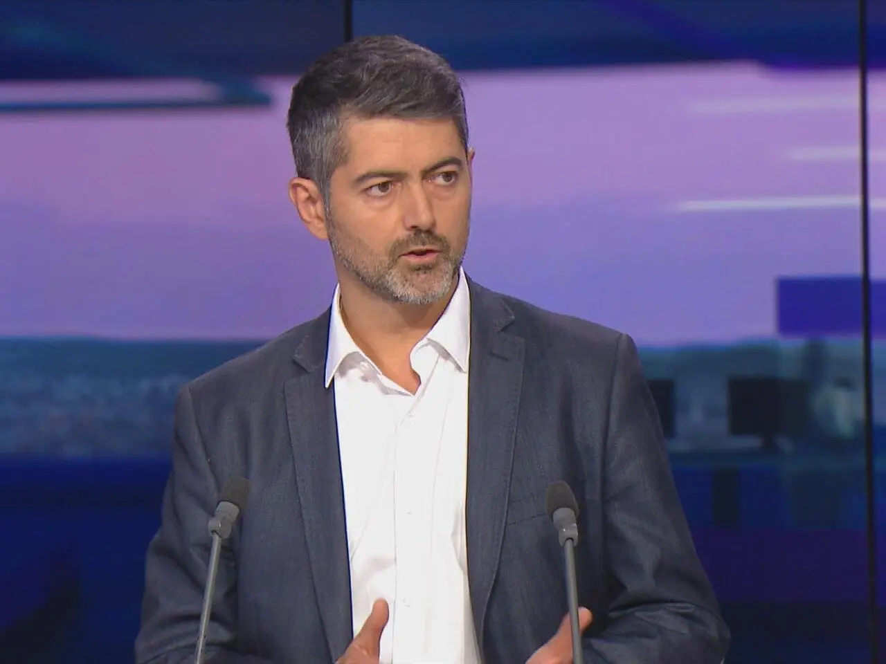 Man with salt-and-pepper hair and beard wearing a dark suit and white shirt speaking at a podium with two microphones.