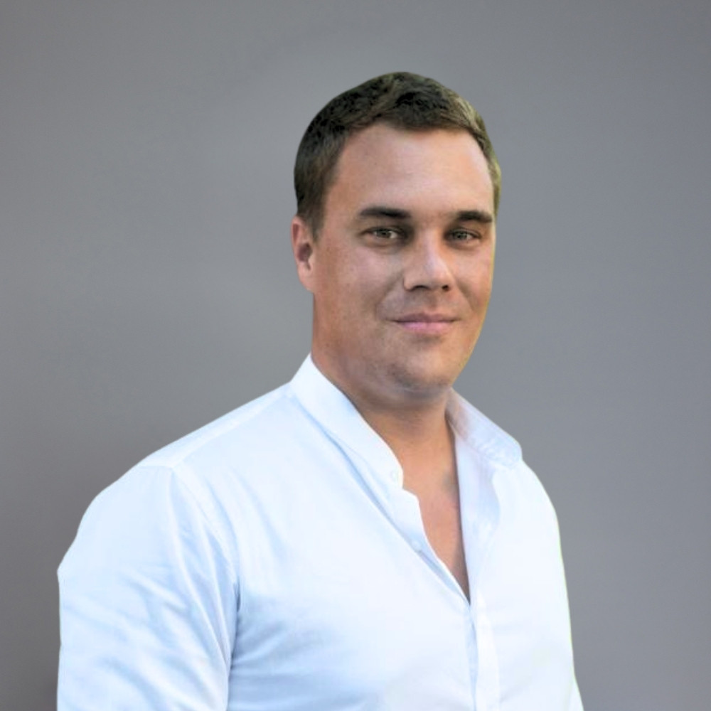 Portrait of a man with short brown hair wearing a white button-up shirt against a plain gray background.